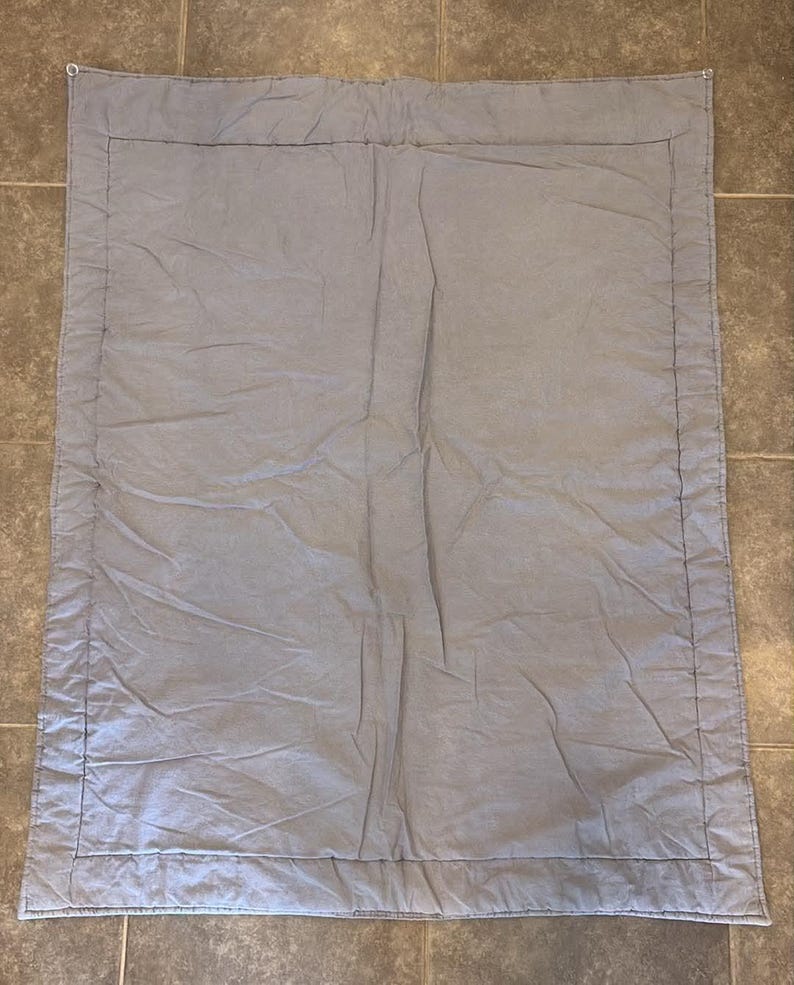 May include: A rectangular, light gray fabric blanket featuring a wide border stitched around the perimeter. The textile has a soft, slightly wrinkled texture and includes small metal grommets at the top two corners for hanging or display purposes.