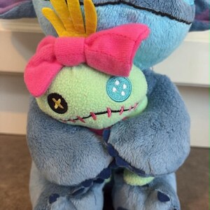 K&ouml;nnte beinhalten: Ein blaues Stitch-Pl&uuml;schtier h&auml;lt eine Scrump-Puppe. Stitch hat gro&szlig;e Ohren, eine dunkelblaue Nase und schwarze Augen. Scrump ist gr&uuml;n mit einer rosa Schleife und einer gelben Krone. Die Spielzeuge befinden sich auf einer braunen Oberfl&auml;che.