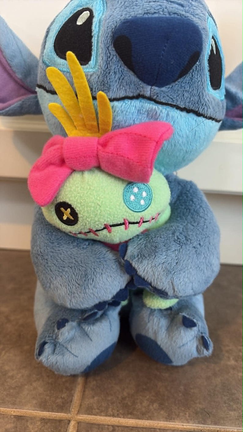 K&ouml;nnte beinhalten: Ein blaues Stitch-Pl&uuml;schtier h&auml;lt eine Scrump-Puppe. Stitch hat gro&szlig;e Ohren, eine dunkelblaue Nase und schwarze Augen. Scrump ist gr&uuml;n mit einer rosa Schleife und einer gelben Krone. Die Spielzeuge befinden sich auf einer braunen Oberfl&auml;che.