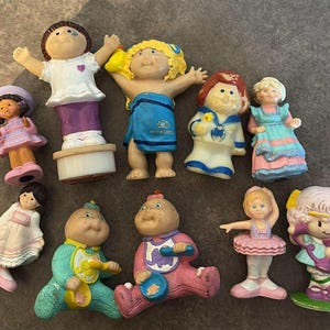 Könnte beinhalten: Eine Sammlung von Vintage-Cabbage-Patch-Kids-Figuren. Die Figuren sind in verschiedenen Posen und Outfits, darunter ein Matrose, eine Ballerina und Babys. Die Farbpalette umfasst Pastelltöne mit Details wie Schleifen, Hüten und Lätzchen.