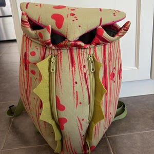 May include: A beige backpack with a red speckled pattern and a top flap resembling a mouth. The bag features two vertical zippered pockets on the front, each flanked by decorative green felt spikes, and adjustable green shoulder straps.