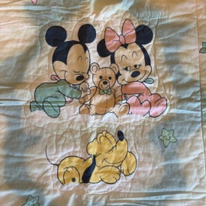 Puede incluir: Una manta acolchada para beb&eacute; de color blanco con un gr&aacute;fico central de Mickey Mouse beb&eacute;, Minnie Mouse beb&eacute; y un osito de peluche, con un perro Pluto amarillo debajo. El fondo est&aacute; decorado con peque&ntilde;as estrellas de colores pastel en rosa, amarillo y verde.