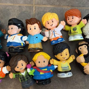 Puede incluir: Una colección de once figuras de juguete de plástico que incluyen varias personas con atuendos coloridos, una oveja blanca y un chimpancé negro que sostiene un plátano amarillo. Una figura lleva una gorra azul con las letras ABC impresas.