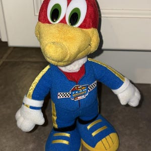 Puede incluir: Mu&ntilde;eco de peluche de Woody Woodpecker con un traje de carreras azul con detalles amarillos. El mu&ntilde;eco tiene la cabeza roja, pico amarillo y ojos verdes grandes. El traje tiene el texto "Team Woody NASCAR".