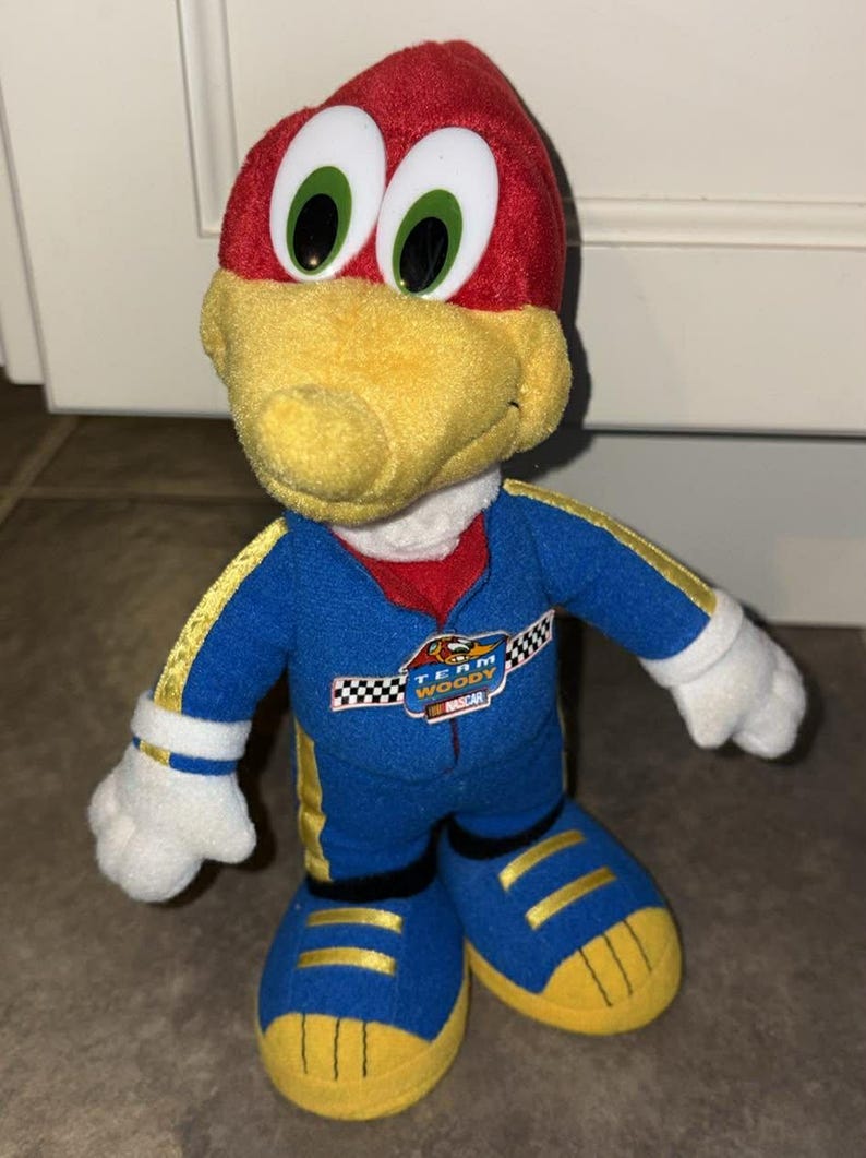 Puede incluir: Mu&ntilde;eco de peluche de Woody Woodpecker con un traje de carreras azul con detalles amarillos. El mu&ntilde;eco tiene la cabeza roja, pico amarillo y ojos verdes grandes. El traje tiene el texto "Team Woody NASCAR".