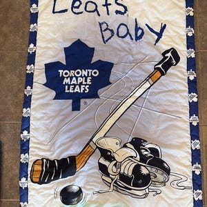 May include: A white baby quilt with a blue border featuring the text "Leafs Baby" and the Toronto Maple Leafs logo. The quilt has a hockey stick, skates, and a puck design.
