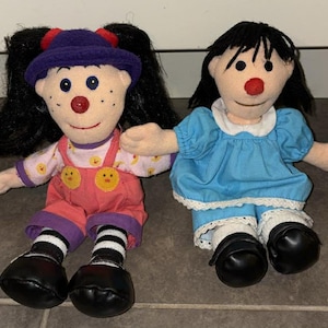 Puede incluir: Dos muñecas de peluche con pelo negro, narices rojas y mejillas rosadas. Una muñeca lleva un sombrero morado, un mono rosa y zapatos negros. La otra muñeca lleva un vestido azul y zapatos negros. Ambas muñecas tienen los brazos extendidos.