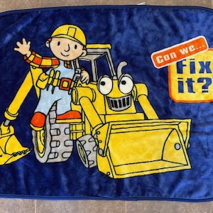 Könnte beinhalten: Eine rechteckige blaue Plüschdecke mit einer Grafik von Bob der Baumeister neben einem gelben Baufahrzeug. Der Text Can we... Fix it? ist in einer gelben und orangefarbenen Sprechblase auf der rechten Seite des weichen Stoffes abgebildet.