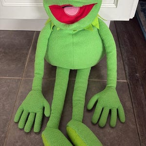 May include: A plush frog toy featuring a bright green fleece body, a wide red mouth with a pink tongue, and large white eyes with black pupils. The character has long, thin arms and legs, a rounded torso, and a pointed green collar around the neck.