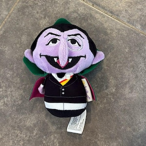 Puede incluir: Un peluche del Conde de Barrio Sésamo. El juguete tiene una cara morada, pelo negro y un traje negro con una capa roja. El juguete tiene una etiqueta adjunta.
