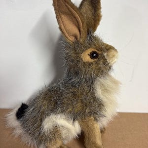 May include: A plush rabbit toy featuring realistic brown, grey, and white faux fur. The figure has long, upright ears with tan inner lining, dark brown eyes, a small nose, and a short tail with a black tip, captured in a seated position.