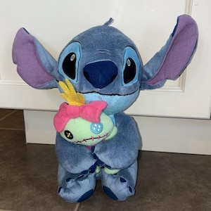 K&ouml;nnte beinhalten: Ein blauer Stitch-Pl&uuml;sch mit gro&szlig;en Ohren und schwarzen Augen, der eine kleine gr&uuml;ne Puppe mit einer rosa Schleife h&auml;lt. Stitch ist eine Figur aus dem Zeichentrickfilm Lilo & Stitch.