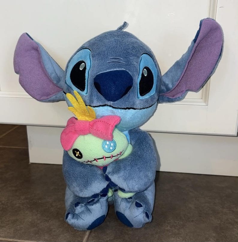 K&ouml;nnte beinhalten: Ein blauer Stitch-Pl&uuml;sch mit gro&szlig;en Ohren und schwarzen Augen, der eine kleine gr&uuml;ne Puppe mit einer rosa Schleife h&auml;lt. Stitch ist eine Figur aus dem Zeichentrickfilm Lilo & Stitch.