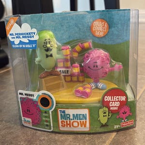 Puede incluir: Un juego de juguetes de Mr. Men Show empaquetado, con las figuras de Mr. Messy y Mr. Pernickety. El embalaje incluye el texto "Collect & Connect Them!" y "Collector Card Inside!" y el logotipo del programa.