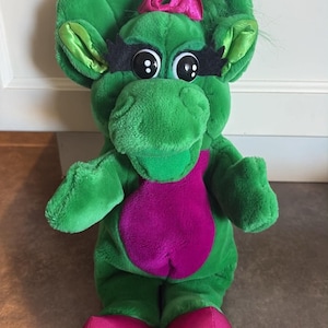 May include: A green plush dinosaur toy with a purple belly, large black eyes with long black eyelashes, and a small pink bow on its head. The dinosaur has shiny pink feet, with the word Bop embroidered in yellow on the right foot.
