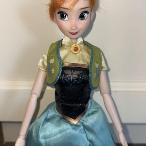 K&ouml;nnte beinhalten: Eine Puppe mit zu einem Dutt frisiertem rotbraunem Haar, Sommersprossen und gr&uuml;nen Augen. Die Puppe tr&auml;gt ein hellgelbes kurz&auml;rmeliges Oberteil, eine gr&uuml;ne Weste mit Blumenmuster, ein schwarzes Mieder und einen langen blauen Rock mit Sonnenblumen.