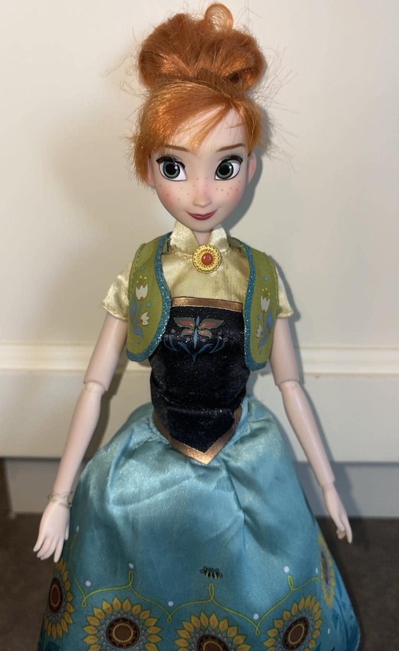 K&ouml;nnte beinhalten: Eine Puppe mit zu einem Dutt frisiertem rotbraunem Haar, Sommersprossen und gr&uuml;nen Augen. Die Puppe tr&auml;gt ein hellgelbes kurz&auml;rmeliges Oberteil, eine gr&uuml;ne Weste mit Blumenmuster, ein schwarzes Mieder und einen langen blauen Rock mit Sonnenblumen.
