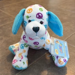 May include: A plush dog toy with white fur featuring a colourful peace sign pattern in orange, purple, green, and blue. The dog has bright blue ears and a black stitched nose. A rectangular tag attached to the side reads Webkinz discover a virtual world.