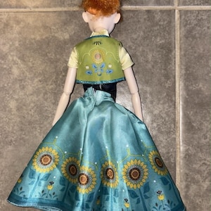 K&ouml;nnte beinhalten: Eine Puppe mit orangefarbenem Haar, das zu einem Dutt hochgesteckt ist, tr&auml;gt ein hellgelbes Kurzarm-Oberteil, eine gr&uuml;ne Weste mit Blumenstickerei und einen langen, blaugr&uuml;nen Rock mit einem Muster aus gelben Sonnenblumen und kleinen blauen Bl&uuml;ten.