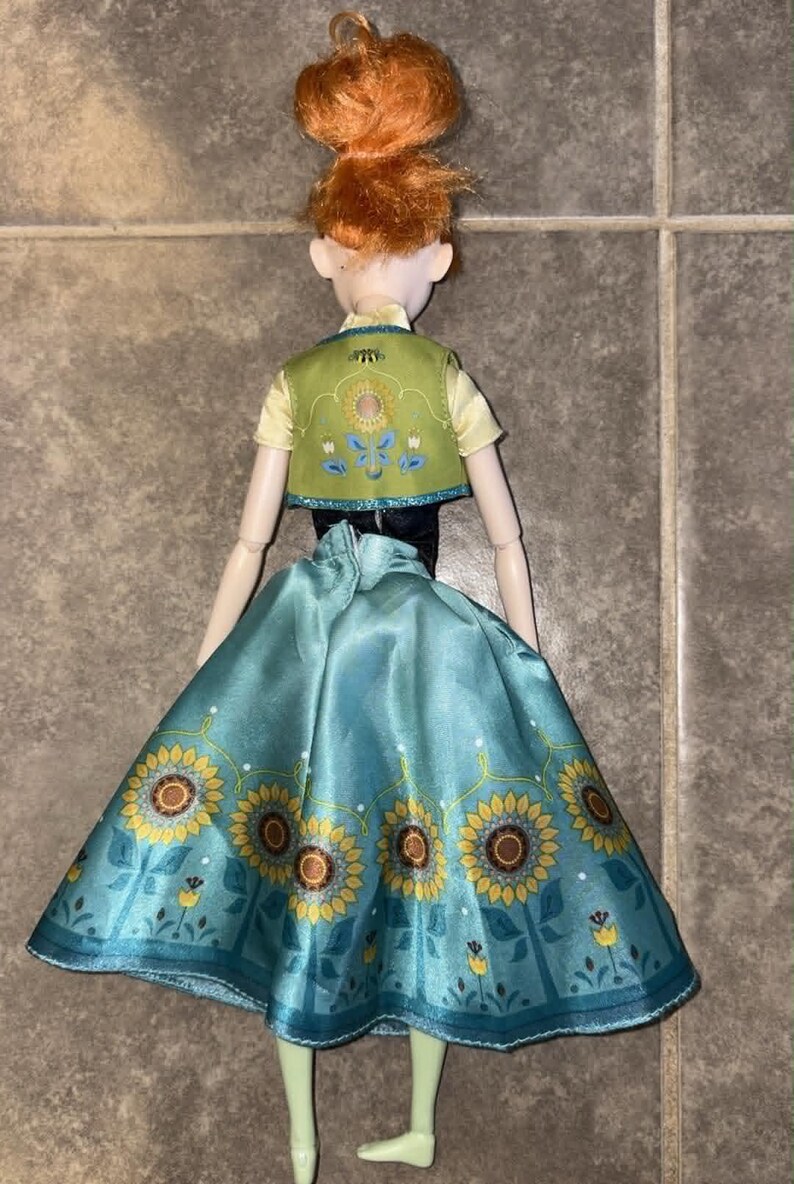 K&ouml;nnte beinhalten: Eine Puppe mit orangefarbenem Haar, das zu einem Dutt hochgesteckt ist, tr&auml;gt ein hellgelbes Kurzarm-Oberteil, eine gr&uuml;ne Weste mit Blumenstickerei und einen langen, blaugr&uuml;nen Rock mit einem Muster aus gelben Sonnenblumen und kleinen blauen Bl&uuml;ten.