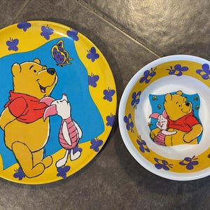 Può includere: Un set di piatti e ciotole gialle con Winnie the Pooh e Piglet. Il piatto ha uno sfondo blu con Winnie the Pooh che tiene Piglet e una farfalla. La ciotola ha un design simile con Winnie the Pooh e Piglet che si abbracciano.