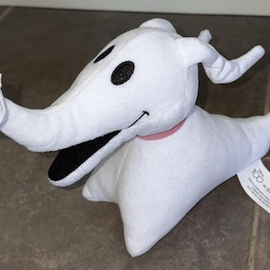 May include: A plush toy of Zero, the ghost dog from The Nightmare Before Christmas. The white dog has black eyes, a long snout, and a pink collar. The plush has a tag attached.