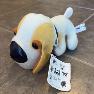 May include: A plush dog toy with a white body, tan ears, and a black nose. The toy has a blue collar and a tag that reads "THE DOG" and features various dog illustrations. The toy is sitting on a brown surface.