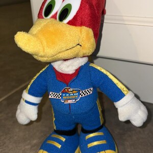 Puede incluir: Mu&ntilde;eco de peluche de Woody Woodpecker con un traje de carreras azul con detalles amarillos y la inscripci&oacute;n "Team Woody NASCAR". El mu&ntilde;eco tiene la cabeza roja, pico amarillo, guantes y zapatos blancos.