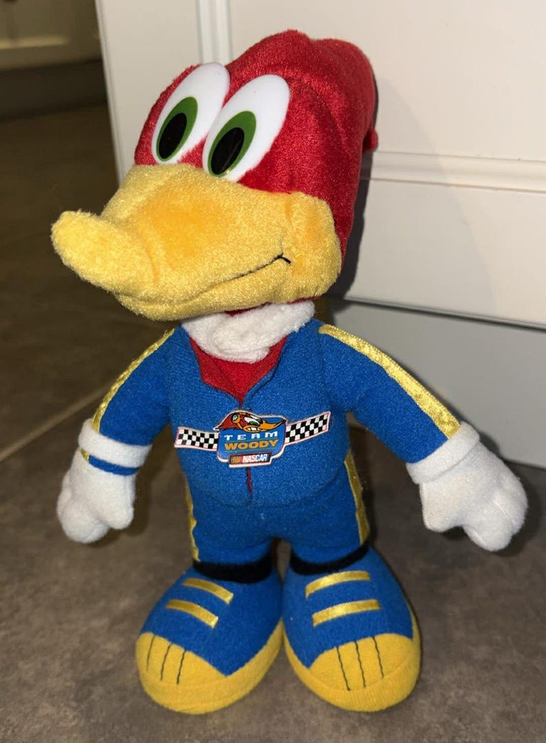 Puede incluir: Mu&ntilde;eco de peluche de Woody Woodpecker con un traje de carreras azul con detalles amarillos y la inscripci&oacute;n "Team Woody NASCAR". El mu&ntilde;eco tiene la cabeza roja, pico amarillo, guantes y zapatos blancos.