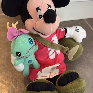 Puede incluir: Un mu&ntilde;eco de peluche de Mickey Mouse con una camisa hawaiana roja con dise&ntilde;os florales blancos, una correa verde y zapatos verdes. Mickey sostiene un peque&ntilde;o mu&ntilde;eco de Stitch turquesa con un lazo rosa. El mu&ntilde;eco tiene orejas negras y una cara rosa.