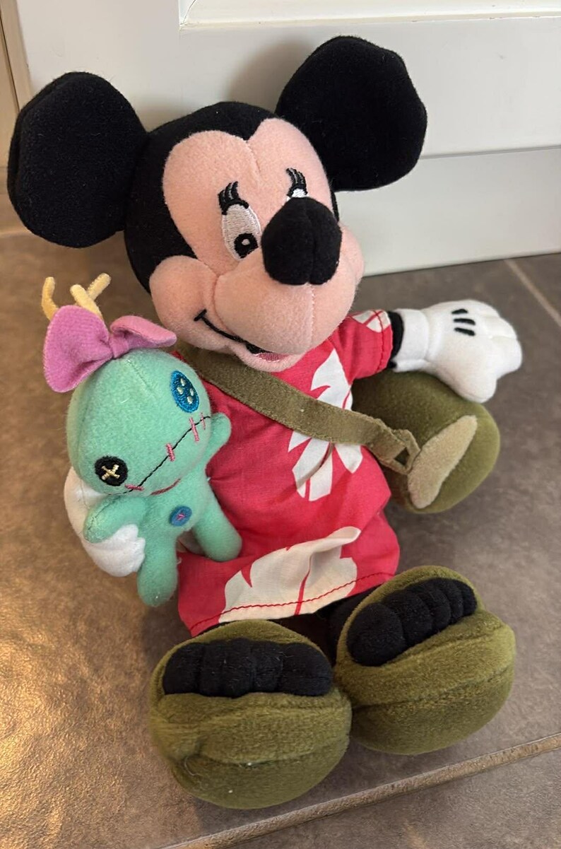 Puede incluir: Un mu&ntilde;eco de peluche de Mickey Mouse con una camisa hawaiana roja con dise&ntilde;os florales blancos, una correa verde y zapatos verdes. Mickey sostiene un peque&ntilde;o mu&ntilde;eco de Stitch turquesa con un lazo rosa. El mu&ntilde;eco tiene orejas negras y una cara rosa.