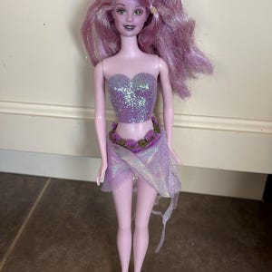 May include: A doll with light purple skin and long, wavy lavender hair featuring subtle glitter accents. The doll wears a strapless, glittery purple bodice, a waist belt adorned with small purple flowers, and a sheer, iridescent purple wrap skirt.