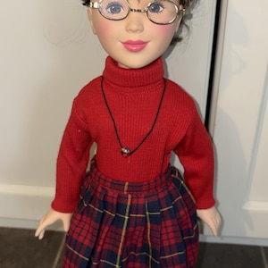 May include: A doll with brown hair and round glasses wearing a red turtleneck sweater, a black cord necklace with a small colourful pendant, and a pleated red and navy blue plaid skirt. The doll has a fair complexion, pink lips, and dark blue eyes.