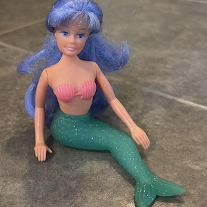 Peut inclure: Une poup&eacute;e sir&egrave;ne avec de longs cheveux bleus ondul&eacute;s, un teint clair et des yeux bleus. La poup&eacute;e porte un haut de bikini rose en forme de coquillage et une queue de sir&egrave;ne verte translucide et paillet&eacute;e avec une texture d'&eacute;cailles.