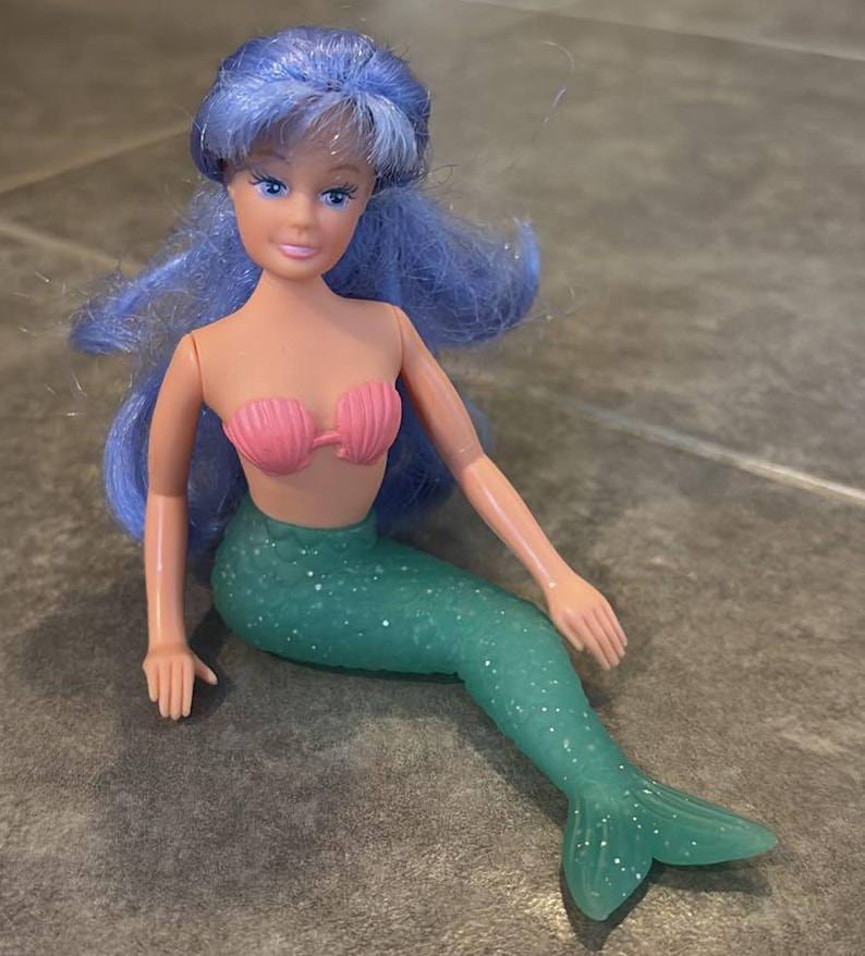 Peut inclure: Une poup&eacute;e sir&egrave;ne avec de longs cheveux bleus ondul&eacute;s, un teint clair et des yeux bleus. La poup&eacute;e porte un haut de bikini rose en forme de coquillage et une queue de sir&egrave;ne verte translucide et paillet&eacute;e avec une texture d'&eacute;cailles.