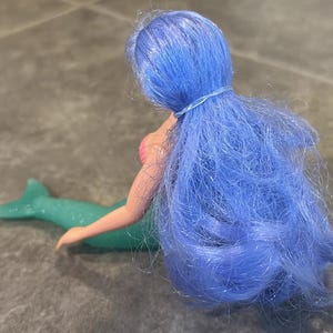 Peut inclure: Une poup&eacute;e sir&egrave;ne aux longs cheveux bleu vif attach&eacute;s par un petit &eacute;lastique. La poup&eacute;e a une peau claire, un haut en forme de coquillage rose et une queue de sir&egrave;ne verte, translucide et scintillante, s'&eacute;tendant horizontalement.