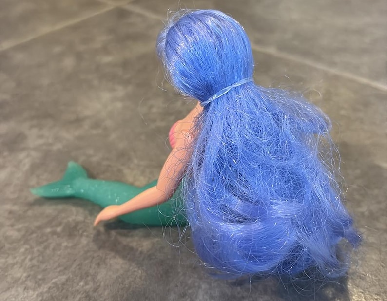 Peut inclure: Une poup&eacute;e sir&egrave;ne aux longs cheveux bleu vif attach&eacute;s par un petit &eacute;lastique. La poup&eacute;e a une peau claire, un haut en forme de coquillage rose et une queue de sir&egrave;ne verte, translucide et scintillante, s'&eacute;tendant horizontalement.