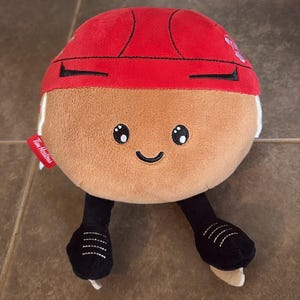 May include: A round, tan plush toy shaped like a Tim Hortons Timbits donut hole, wearing a red hockey helmet and black ice skates. The plush features embroidered black eyes and a smile, with a small red fabric tag labeled Tim Hortons on the side.