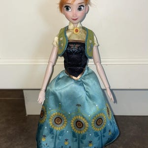 K&ouml;nnte beinhalten: Eine Puppe mit zu einem Dutt frisiertem rotbraunem Haar, bekleidet mit einem hellgelben Kurzarm-Oberteil, einem schwarzen Mieder, einer gr&uuml;nen Weste mit Blumenakzenten und einem hellblauen Rock mit Sonnenblumenmuster und kleinen gelben Bl&uuml;ten.