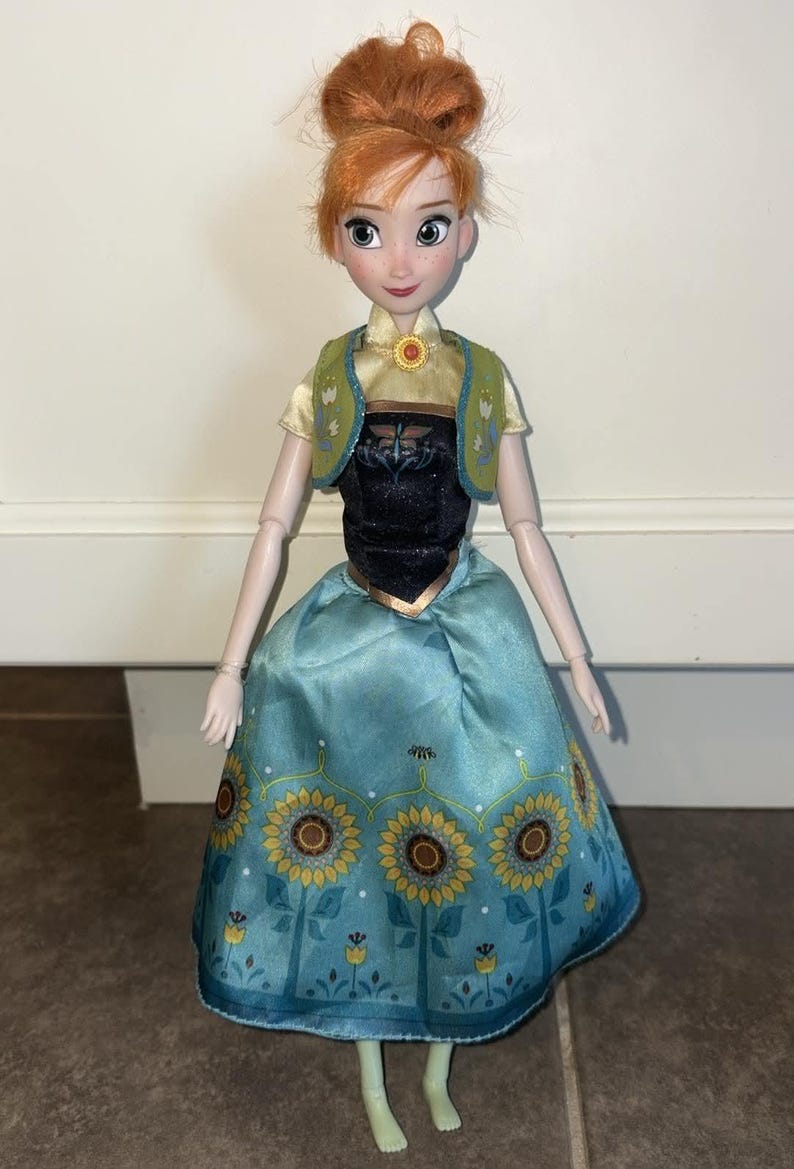 K&ouml;nnte beinhalten: Eine Puppe mit zu einem Dutt frisiertem rotbraunem Haar, bekleidet mit einem hellgelben Kurzarm-Oberteil, einem schwarzen Mieder, einer gr&uuml;nen Weste mit Blumenakzenten und einem hellblauen Rock mit Sonnenblumenmuster und kleinen gelben Bl&uuml;ten.