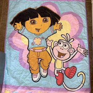 Puede incluir: Un edredón con el tema de Dora la Exploradora con Dora y Botas el mono. El edredón tiene un fondo azul claro con un gran diseño de flores en el centro. Dora lleva una camisa azul y pantalones naranjas. Botas lleva botas rojas.