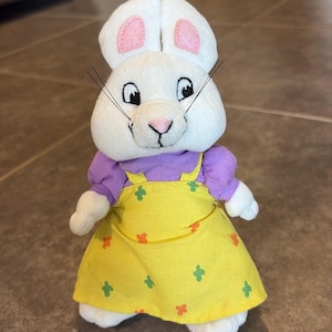 May include: A plush white rabbit toy with embroidered facial features, black whiskers, and pink inner ears. The rabbit wears a purple short-sleeved shirt under a yellow jumper dress featuring a small repeating pattern of orange and green cross shapes.