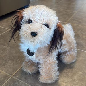 以下が含まれることがあります： クリーム色の体と濃い茶色のアクセントが耳にある犬のぬいぐるみ。犬は黒い鼻と目、黒い首輪をしています。おもちゃはタイルの上に立っています。