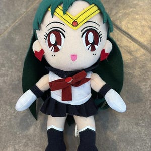 May include: A plush doll of a character with green hair, a gold tiara, and red eyes. The doll wears a white top with a dark red bow, a navy blue skirt, and navy blue boots. The doll has white gloves and red earrings.