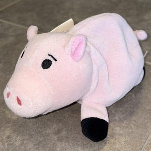 Puede incluir: Un peluche rosa con forma de cerdo con ojos, orejas y pezu&ntilde;as negras. El cerdo tiene una peque&ntilde;a cola y una etiqueta adjunta. El juguete est&aacute; hecho de material suave y est&aacute; dise&ntilde;ado para parecerse a un cerdo.
