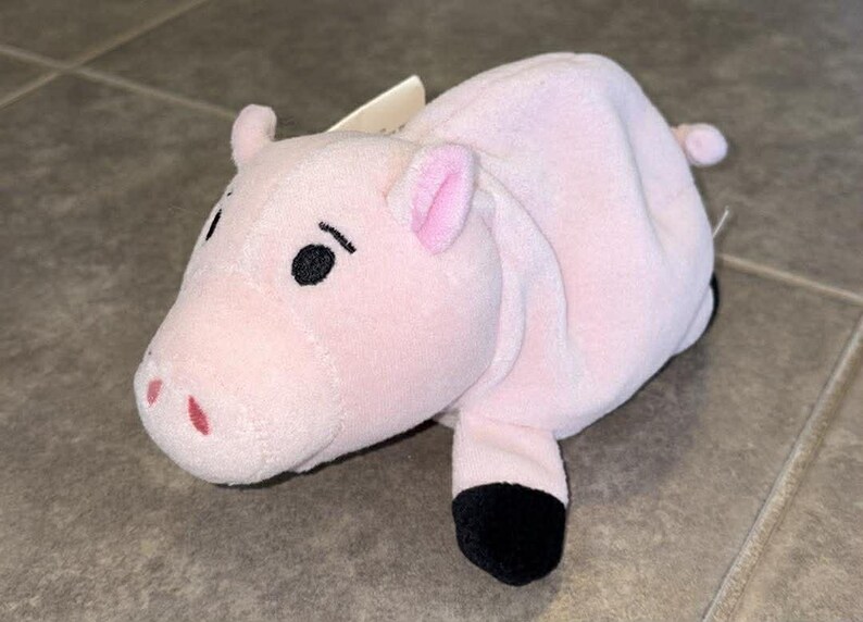Puede incluir: Un peluche rosa con forma de cerdo con ojos, orejas y pezu&ntilde;as negras. El cerdo tiene una peque&ntilde;a cola y una etiqueta adjunta. El juguete est&aacute; hecho de material suave y est&aacute; dise&ntilde;ado para parecerse a un cerdo.