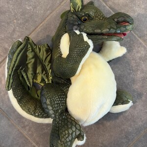 Pode incluir: Um dragão de pelúcia com um exterior texturizado de escamas verde-escuras, uma barriga cor de creme e asas brilhantes verde-oliva. A criatura tem uma cabeça detalhada com olhos visíveis e uma boca ligeiramente aberta com interior vermelho.