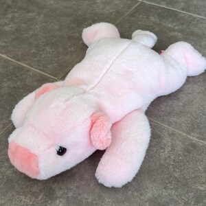 Puede incluir: Un peluche de cerdo de color rosa claro con pelaje suave, orejas pequeñas y redondeadas, un hocico y ojos oscuros. El animal de peluche está en posición tumbada con las cuatro patas extendidas y tiene una pequeña cola en la parte trasera.