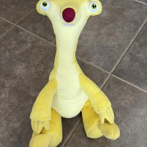 Pode incluir: Um brinquedo de pelúcia amarelo em forma de bicho-preguiça com um pescoço longo, um nariz vermelho escuro e olhos verdes grandes. O personagem tem uma cabeça larga com órbitas salientes, braços longos e pernas curtas. O tecido é macio.