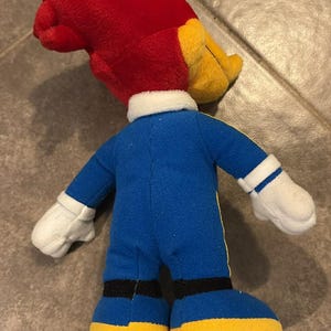 Puede incluir: Un peluche de un personaje de dibujos animados, un p&aacute;jaro carpintero, visto desde atr&aacute;s. El juguete tiene una cabeza roja, un pico amarillo y un mono azul con detalles amarillos. Guantes blancos y zapatos amarillos completan el atuendo. El juguete mide unos 25 cm de alto.
